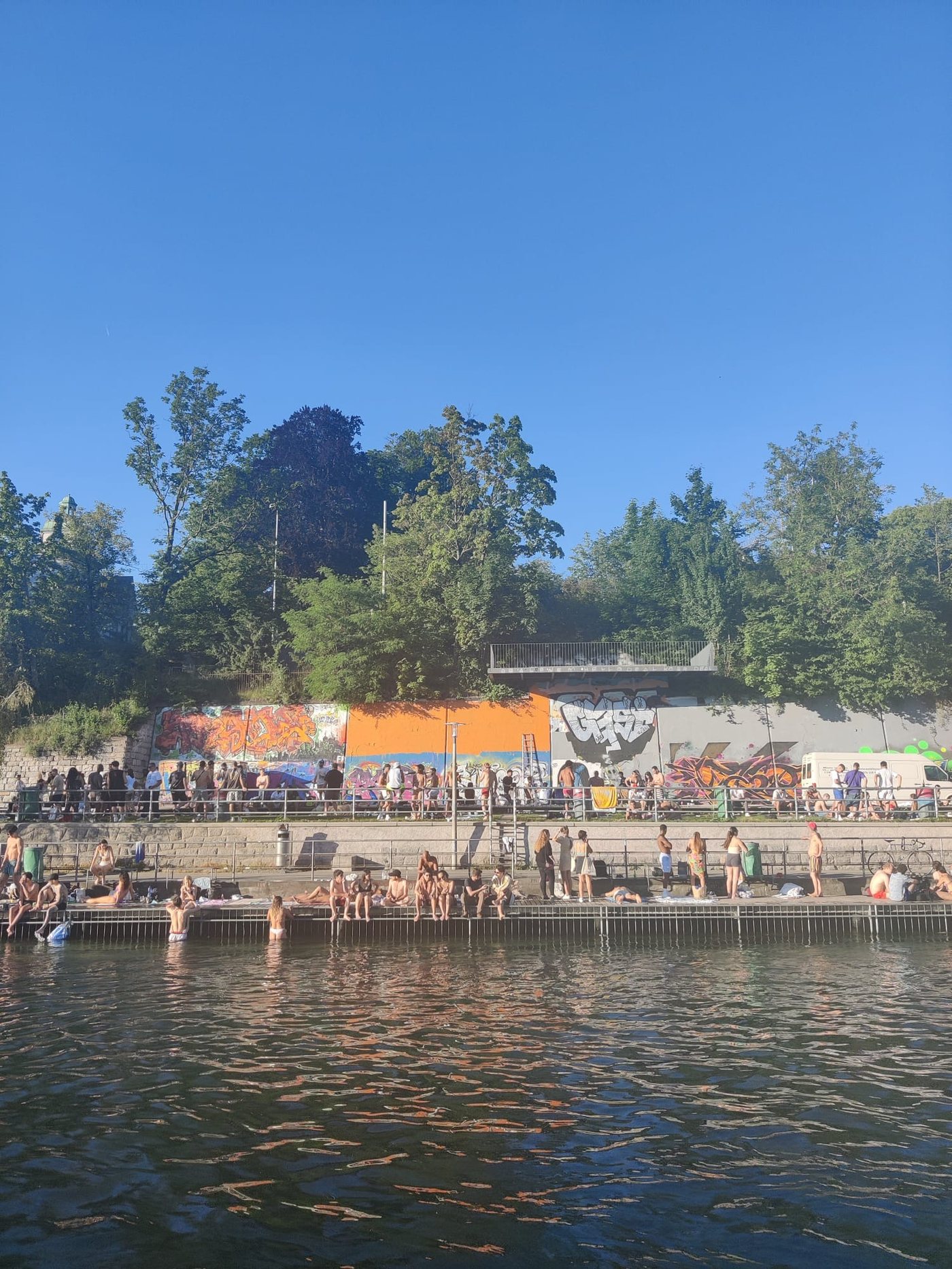 People gathered beside a river with graffiti walls and trees