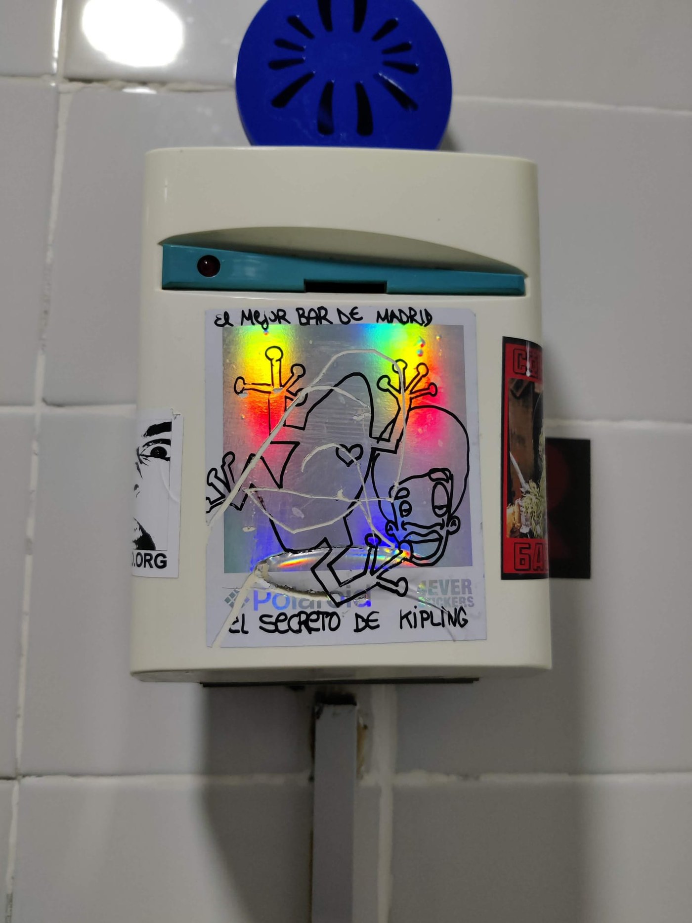 A personal photograph of a sticker-covered wall-mounted dispenser